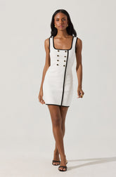 A woman is wearing a sleeveless, fitted white dress with black trim along the square neckline, armholes, and a vertical line running from the neckline to the hem. The dress features two rows of gold buttons on the front, arranged in a double-breasted style. She completes her outfit with black open-toe high-heeled sandals and gold hoop earrings.